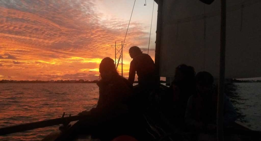 the silhouettes of two people on a sailboat appear before an orange sky while the sun sets. 